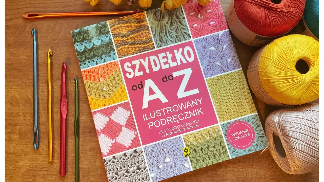 Książka "Szydełko od A do Z" z kolorowymi motywami, obok niej kilka szydełek, w tym jedno długie, oraz kłębki włóczki.