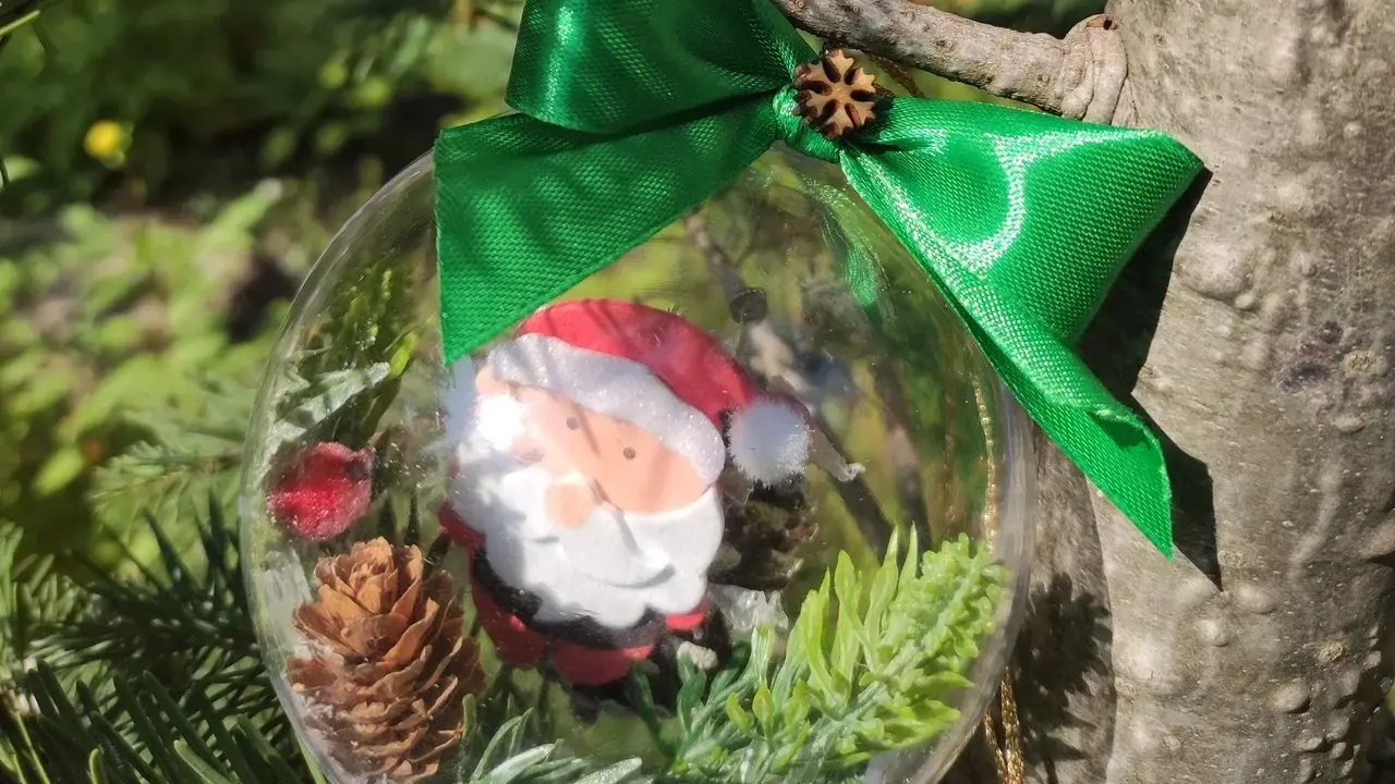 Przezroczysta bombka DIY z Mikołajem, szyszką i gałązkami, przewiązana zieloną wstążką, wisi na choince.