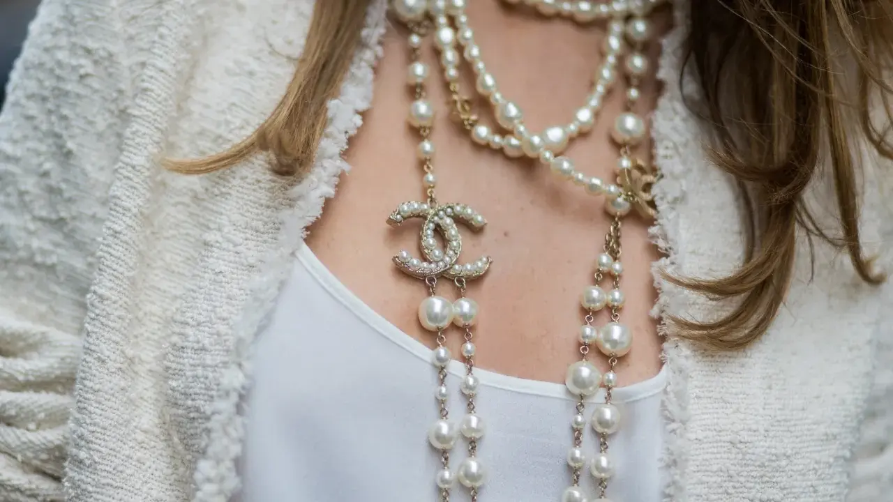 Elegancka kobieta w białej marynarce i białej bluzce, ozdobiona wieloma naszyjnikami z pereł i logo Chanel. Idealny wybór dla kogo perły.