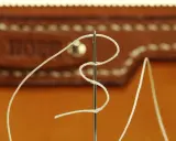 Zbliżenie na igłę do szycia ręcznego z nawleczoną białą nicią, na tle brązowej skóry z ozdobnymi przeszyciami. Różne rodzaje igieł do szycia ręcznego mogą mieć różne zastosowania.