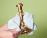 Dłoń czyści mosiężny świecznik jasnoniebieską ściereczką na zielonym tle.