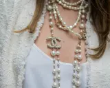 Elegancka kobieta w białej marynarce i białej bluzce, ozdobiona wieloma naszyjnikami z pereł i logo Chanel. Idealny wybór dla kogo perły.