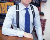 Mężczyzna w błękitnej koszuli, granatowych szelkach i krawacie z ptakami.
