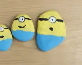 Urocze malowanie kamieni w kształcie Minionków. Trzy kamienie, każdy z żółtą górą, niebieskimi spodenkami i jednym okiem.