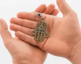 Dłonie trzymające ozdobny amulet w kształcie ręki Fatimy z niebieskim okiem.