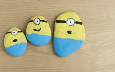 Urocze malowanie kamieni w kształcie Minionków. Trzy kamienie, każdy z żółtą górą, niebieskimi spodenkami i jednym okiem.