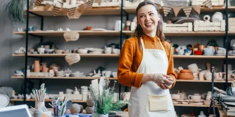 Uśmiechnięta artystka w fartuchu, otoczona ceramiką, zastanawia się, jak wycenić rękodzieło.