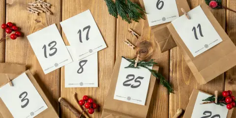 Jak zrobić kalendarz adwentowy z papieru: brązowe torebki z numerami dni, gałązki i czerwone ozdoby na drewnianym tle.