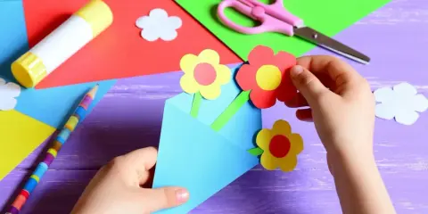 Dziecko tworzy kartkę z papieru w kształcie koperty z kwiatkami, nożyczki, klej i kredki.