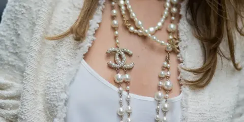 Elegancka kobieta w białej marynarce i białej bluzce, ozdobiona wieloma naszyjnikami z pereł i logo Chanel. Idealny wybór dla kogo perły.