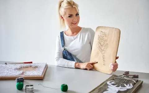 Kobieta prezentuje obraz z nici i gwoździ szablon. Na stole widać inne prace i materiały do tworzenia.