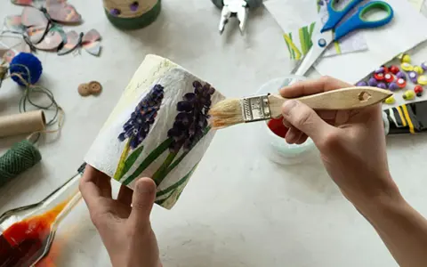 Ręce malują pędzlem ozdobiony kwiatami pojemnik, wokół materiały do decoupage.