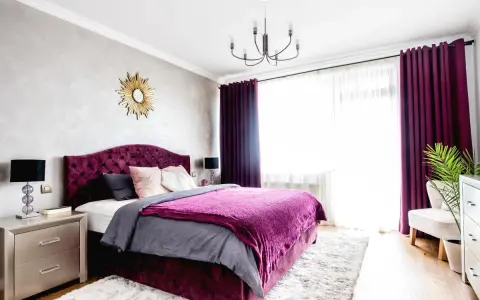 Elegancka sypialnia z łóżkiem w kolorze biskupi, aksamitnym zagłówkiem i narzutą. Obok szafka nocna, lampa i złote lustro.