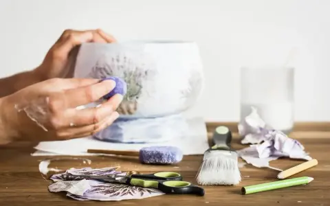 Ręce ozdabiające wazon techniką decoupage. Na stole nożyczki, pędzel, serwetki z lawendą i klej.