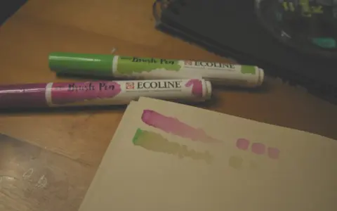 Dwa pisaki Ecoline (zielony i fioletowy) na drewnianym stole, obok próbki kolorów na papierze.