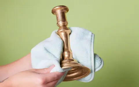 Dłoń czyści mosiężny świecznik jasnoniebieską ściereczką na zielonym tle.
