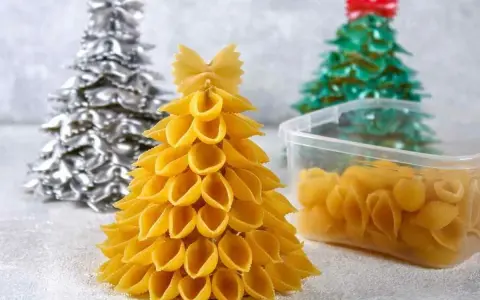 Świąteczne DIY dla dzieci: choinki z makaronu. Złota, srebrna i zielona choinka z różnych rodzajów makaronu, obok pojemnik z muszelkami.