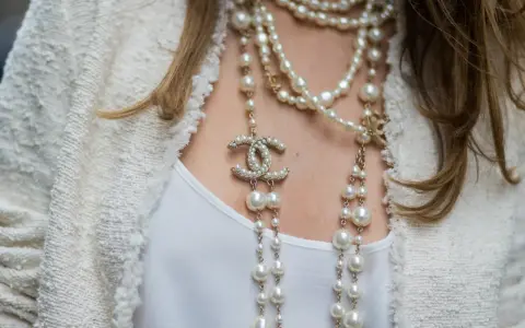 Elegancka kobieta w białej marynarce i białej bluzce, ozdobiona wieloma naszyjnikami z pereł i logo Chanel. Idealny wybór dla kogo perły.