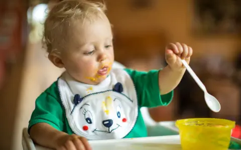 Małe dziecko z marchewkowym puree na buzi i śliniaczku próbuje samodzielnie jeść. Oto jak sprać marchewkę z ubranka!