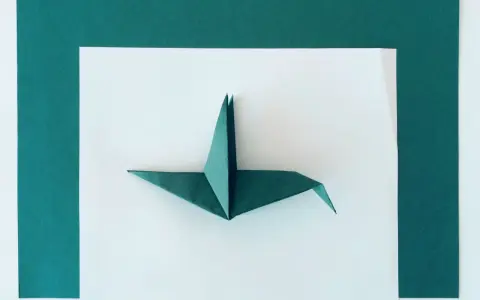Zielony żuraw z papieru, przykład sztuki origami dla dorosłych, leży na białym tle otoczonym ciemnozielonymi ramkami.