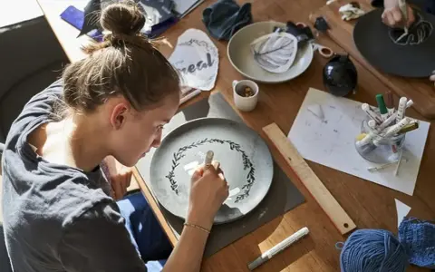 Dziewczyna maluje na talerzu, tworząc wzór. Wokół materiały do rękodzieła, jak do zimna porcelana przepis.