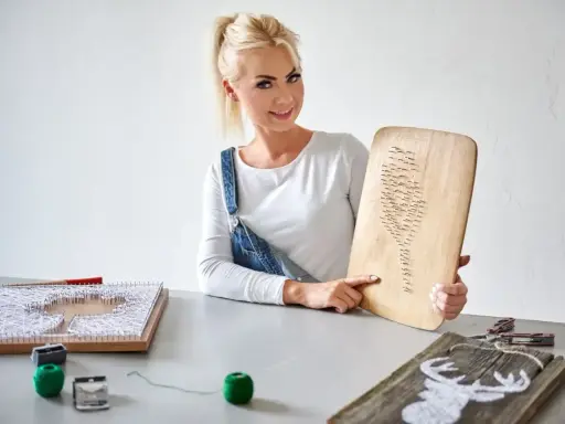Kobieta prezentuje obraz z nici i gwoździ szablon. Na stole widać inne prace i materiały do tworzenia.
