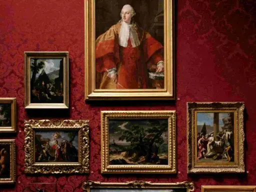 Obrazy w bogatym, malarskim stylu zdobią czerwoną, wzorzystą ścianę. Portret mężczyzny w stroju z epoki, bukiet kwiatów i sceny rodzajowe tworzą galerię.