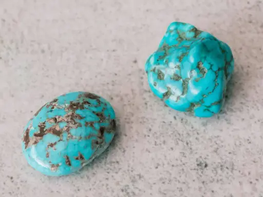 Dwa surowe kamienie turkusowe o niebiesko-zielonym kolorze i brązowych żyłkach, leżące na szarej, chropowatej powierzchni.