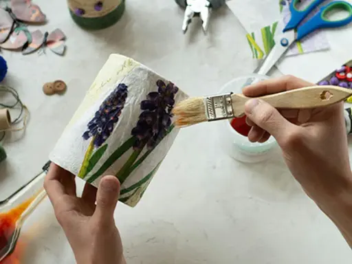 Ręce malują pędzlem ozdobiony kwiatami pojemnik, wokół materiały do decoupage.