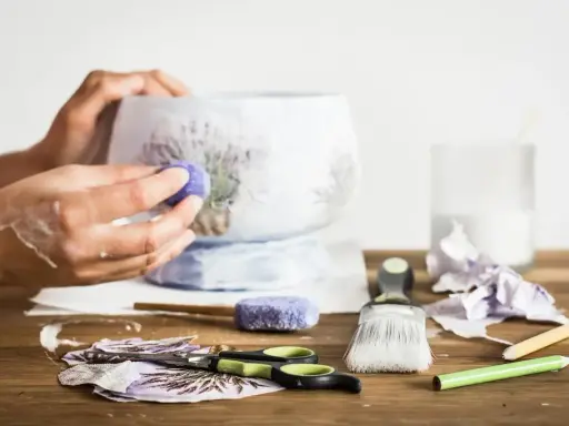 Ręce ozdabiające wazon techniką decoupage. Na stole nożyczki, pędzel, serwetki z lawendą i klej.