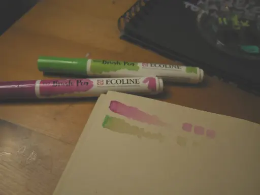 Dwa pisaki Ecoline (zielony i fioletowy) na drewnianym stole, obok próbki kolorów na papierze.