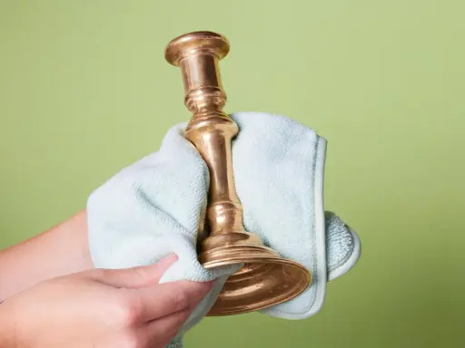 Dłoń czyści mosiężny świecznik jasnoniebieską ściereczką na zielonym tle.