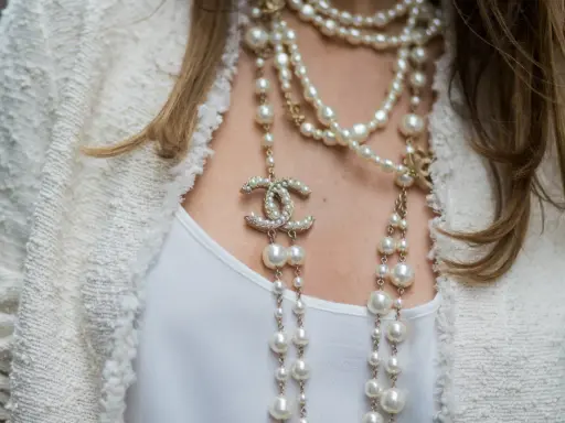 Elegancka kobieta w białej marynarce i białej bluzce, ozdobiona wieloma naszyjnikami z pereł i logo Chanel. Idealny wybór dla kogo perły.