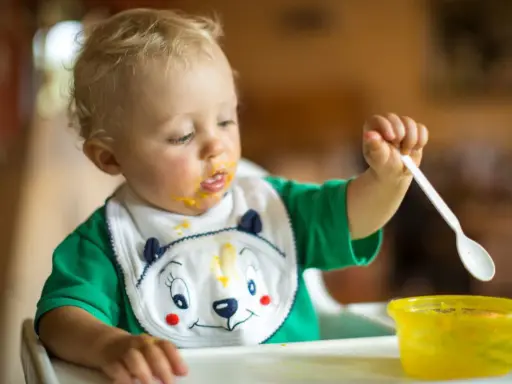 Małe dziecko z marchewkowym puree na buzi i śliniaczku próbuje samodzielnie jeść. Oto jak sprać marchewkę z ubranka!