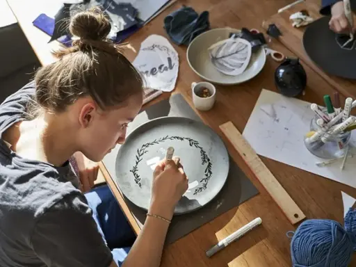 Dziewczyna maluje na talerzu, tworząc wzór. Wokół materiały do rękodzieła, jak do zimna porcelana przepis.