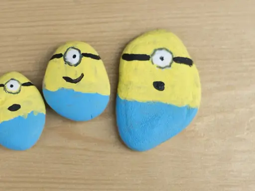 Urocze malowanie kamieni w kształcie Minionków. Trzy kamienie, każdy z żółtą górą, niebieskimi spodenkami i jednym okiem.