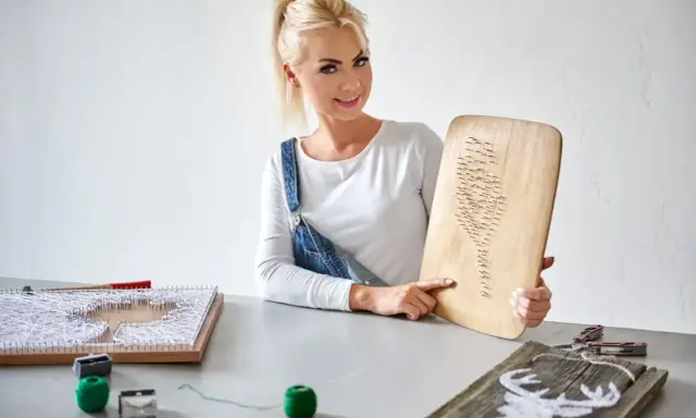 Kobieta prezentuje obraz z nici i gwoździ szablon. Na stole widać inne prace i materiały do tworzenia.