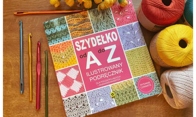 Książka "Szydełko od A do Z" z kolorowymi motywami, obok niej kilka szydełek, w tym jedno długie, oraz kłębki włóczki.