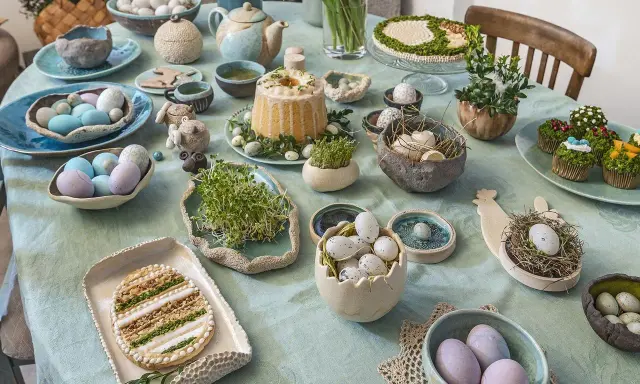 Stół wielkanocny z dekoracjami: pisanki, babeczki, ciasto, ciasteczko w kształcie jajka i kiełki.