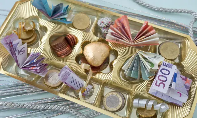 Złote pudełko po czekoladkach wypełnione banknotami euro i monetami.