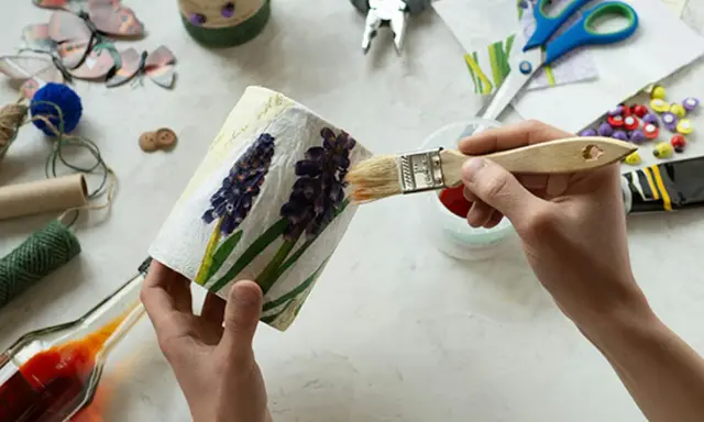 Ręce malują pędzlem ozdobiony kwiatami pojemnik, wokół materiały do decoupage.