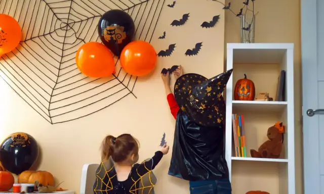 Dzieci w strojach na Halloween dekorują pokój, wieszając nietoperze na ścianie z wielką pajęczyną.