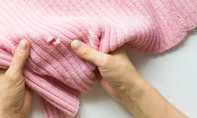 Dłonie trzymają różowy sweter z dziurą, pokazując problem, jak zaszyć dziurę w dresach.