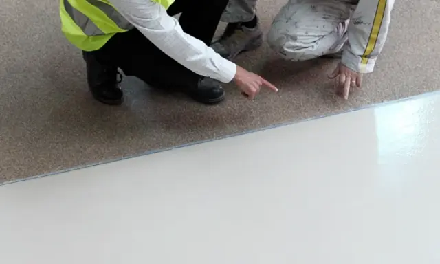 Dwóch pracowników sprawdza, do czego nie przywiera żywica epoksydowa. Jedna osoba w kamizelce odblaskowej wskazuje palcem na granicę między podłożem a nową posadzką.