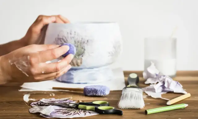 Ręce ozdabiające wazon techniką decoupage. Na stole nożyczki, pędzel, serwetki z lawendą i klej.