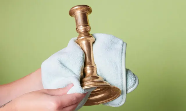 Dłoń czyści mosiężny świecznik jasnoniebieską ściereczką na zielonym tle.
