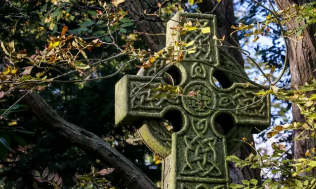 Kamienny krzyż celtycki z misternymi wzorami, otoczony jesiennymi liśćmi. Jego znaczenie symbolizuje wieczność i połączenie.