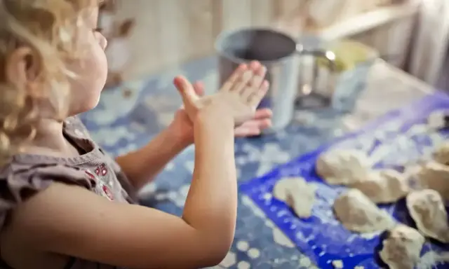 Mała dziewczynka z kręconymi włosami tworzy coś z masy plastycznej. To świetne kreatywne zabawy z 6 latkiem, rozwijające wyobraźnię i zdolności manualne.
