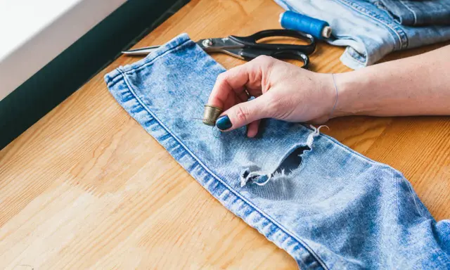 Ręka z naparstkiem i igłą przygotowuje się do naprawy dziury w dżinsach. Widać nożyczki i nici, co sugeruje, jak zaszyć dziurę w cienkim materiale.