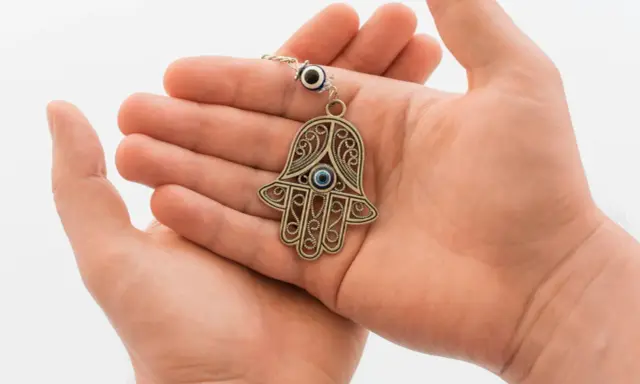 Dłonie trzymające ozdobny amulet w kształcie ręki Fatimy z niebieskim okiem.